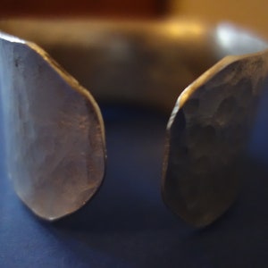 May include: A silver hammered metal cuff bracelet with an open design. The bracelet is made of thick metal and has a rough, textured surface.