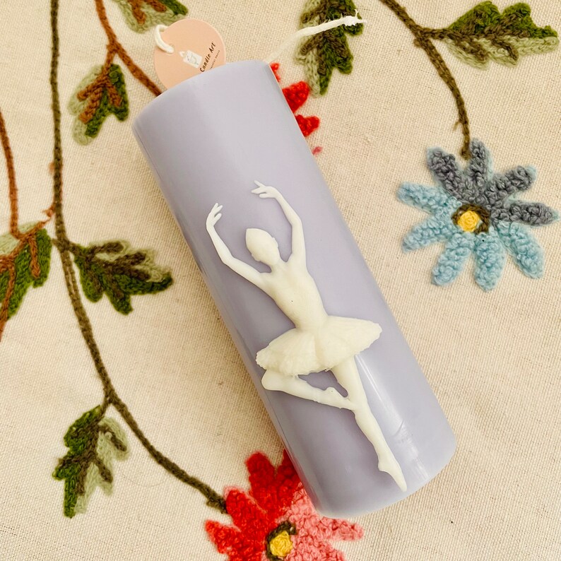 May include: A lavender candle with a white ballerina figurine on the front. The candle has a label that says "Candle Art".