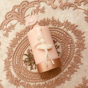 May include: A pink pillar candle with a white ballerina sculpted on the side. The candle has a gold band around the bottom. The candle is on a white lace tablecloth.