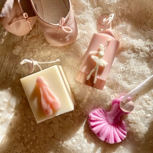 May include: Three pink and white candles shaped like ballet shoes and a ballerina. The ballerina candle is in a pink and gold container. The candles are on a white lace background.