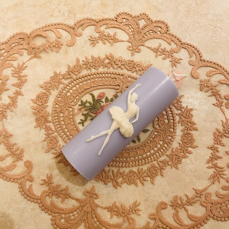 May include: A lavender candle with a white ballerina figurine on the side. The candle is on a lace tablecloth.
