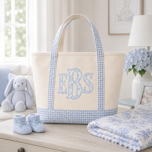 May include: A cream-coloured canvas tote bag featuring blue and white gingham trim on the handles and base. The centre of the bag displays a large, embroidered monogram with the letters EBS in a blue floral pattern on a light background.