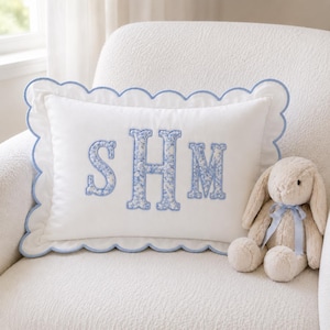 May include: A rectangular white throw pillow with a scalloped edge trimmed in light blue. The center features the embroidered monogram letters SHM in a blue and white floral pattern. Beside the pillow sits a beige plush rabbit toy.