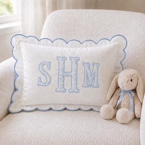 May include: A rectangular white throw pillow with a scalloped edge trimmed in light blue piping. The center features the embroidered letters SHM in a light blue floral pattern. Beside the pillow sits a plush beige rabbit with a light blue ribbon.