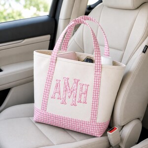 May include: A cream-colored canvas tote bag featuring a pink and white gingham pattern on the base and handles. The front of the bag is embroidered with the letters AMH in a pink floral print. The interior contains a wallet and a white bottle.