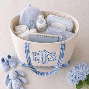 May include: A cream-colored canvas tote bag with blue gingham trim and handles, featuring the embroidered monogram EBS in blue. The bag contains a light blue knit hat with a bear, a baby bottle, a blue gingham pouch, a notebook, and a blue towel.