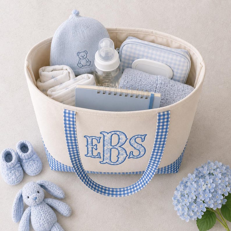 May include: A cream-colored canvas tote bag with blue gingham trim and handles, featuring the embroidered monogram EBS in blue. The bag contains a light blue knit hat with a bear, a baby bottle, a blue gingham pouch, a notebook, and a blue towel.