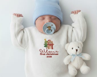 Op de afbeelding: Een pasgeboren baby draagt een lichtblauwe muts en een witte trui met een kerstberenafbeelding en de tekst "Wilson's first christmas 2025". Een witte teddybeer met een blauwe strik ligt ernaast.