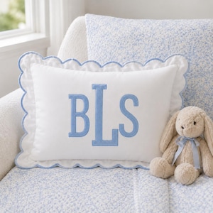 May include: A rectangular white throw pillow featuring a scalloped edge trimmed in light blue piping. The center of the pillow displays a large, embroidered monogram with the letters B, L, and S in a light blue, block-style font.