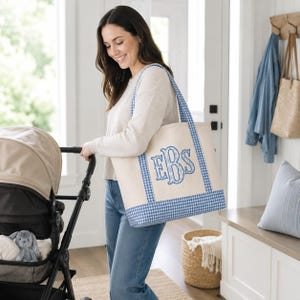 May include: A large off-white canvas tote bag featuring a blue and white gingham check pattern on the base and straps. The center of the bag displays a large, decorative embroidered monogram with the letters EBS in a blue floral print.