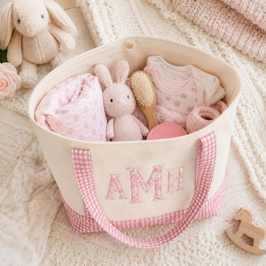 May include: A cream-coloured canvas tote bag featuring pink gingham handles and a matching base. The front is embroidered with the letters AMH in a floral pink fabric. Inside the bag are a pink floral blanket, a crochet bunny, a wooden brush, and clothing.