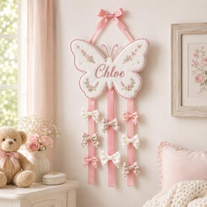 May include: A white fabric butterfly wall hanging featuring the embroidered name Chloe in pink script, accented with small floral patterns. Three long pink ribbons hang from the butterfly, each holding several small decorative hair bows.