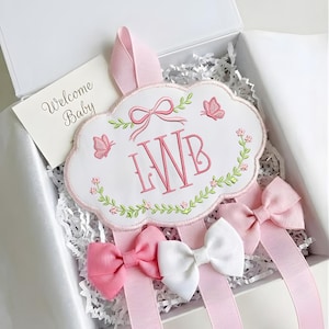 Könnte beinhalten: Ein weißes, wolkenförmiges Baby-Andenken mit rosa Stickerei, Schmetterlingen und den Initialen "LWB". Das Andenken befindet sich in einer weißen Box mit rosa Bändern und Schleifen. Eine Karte mit den Worten "Welcome Baby" ist ebenfalls enthalten.