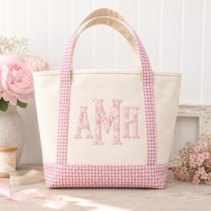 May include: A cream-coloured canvas tote bag featuring pink and white gingham trim on the handles and base. The centre of the bag displays a large, embroidered monogram with the letters AMH in a pink floral pattern against the off-white fabric.