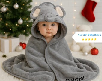 Peut inclure: Un bébé enveloppé dans une serviette à capuche grise avec un motif de visage d'éléphant. Le nom "Gabriel" est brodé sur la serviette. En arrière-plan, un sapin de Noël, une chaussette rouge et un ours en peluche blanc.