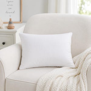 May include: A rectangular white throw pillow with a smooth, solid-colored fabric cover. The pillow is positioned centrally on a light-colored upholstered armchair, featuring a clean, plain surface without any patterns, textures, or decorative embellishments.