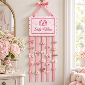 May include: A rectangular wall-hanging hair clip organiser with scalloped edges, featuring a pink ribbon hanger and five vertical pink fabric strips. The top panel displays the monogram EWM and the name Emily William in pink embroidery.