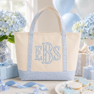 May include: A cream-coloured canvas tote bag featuring light blue and white gingham check trim on the base and handles. The centre of the bag displays a large, embroidered monogram with the letters EBS in a decorative light blue floral pattern.