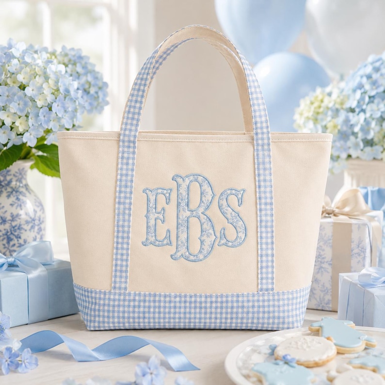 May include: A cream-coloured canvas tote bag featuring light blue and white gingham check trim on the base and handles. The centre of the bag displays a large, embroidered monogram with the letters EBS in a decorative light blue floral pattern.