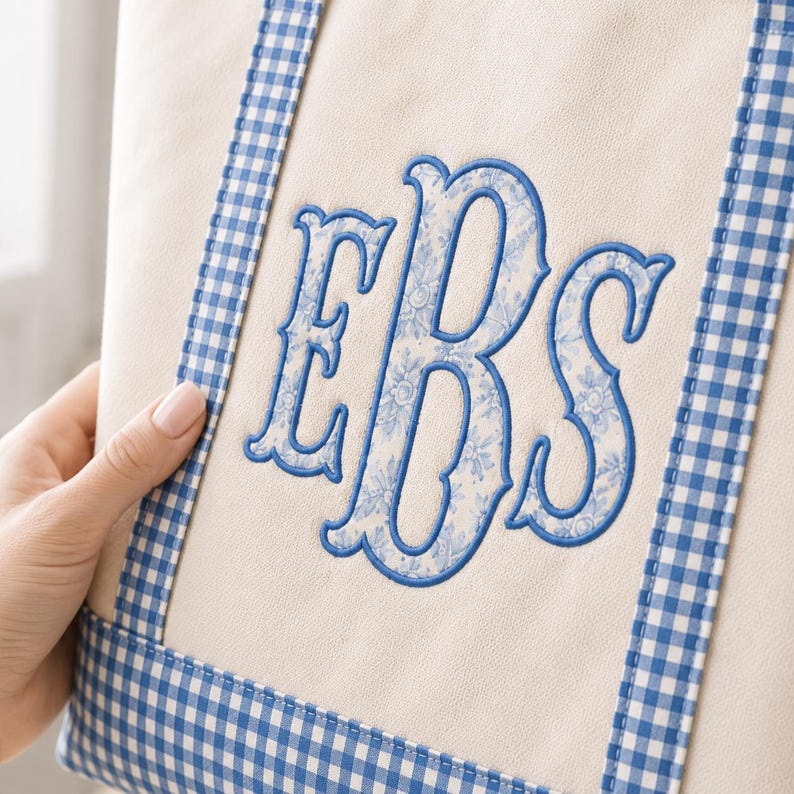 May include: A beige canvas tote bag featuring blue and white gingham trim along the edges. The center of the bag displays a large, embroidered monogram with the letters EBS, filled with a light blue and white floral pattern and outlined in blue.