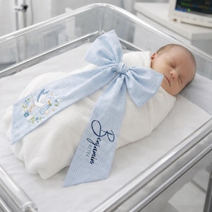 Puede incluir: Un bebé recién nacido envuelto en una manta blanca, adornado con un lazo de cuadros vichy azul claro. El lazo presenta detalles bordados de una cigüeña que lleva un bebé y el nombre "Benjamin Peter" en escritura azul marino.