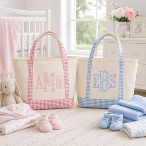 May include: Two canvas tote bags with gingham trim and embroidered monograms. One bag features pink gingham trim and the letters AMH in pink floral embroidery. The other bag features blue gingham trim and the letters EBS in blue floral embroidery.