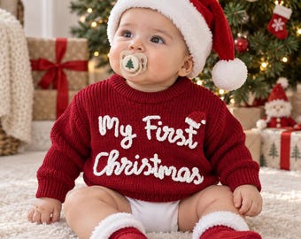 May include: A baby wearing a red knitted sweater with white text reading My First Christmas, a red Santa hat with a white pom-pom, and red socks with white cuffs. The baby has a pacifier with a small green tree graphic in the center.