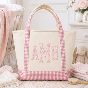May include: A cream-coloured canvas tote bag featuring pink and white gingham trim on the handles and base. The centre of the bag displays the embroidered monogram letters AMH in a pink floral pattern against the neutral fabric background.