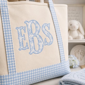 May include: A cream-coloured canvas tote bag featuring light blue gingham trim on the handles and base. The centre of the bag displays a large, embroidered monogram with the letters EBS in a light blue floral pattern with a solid blue border.