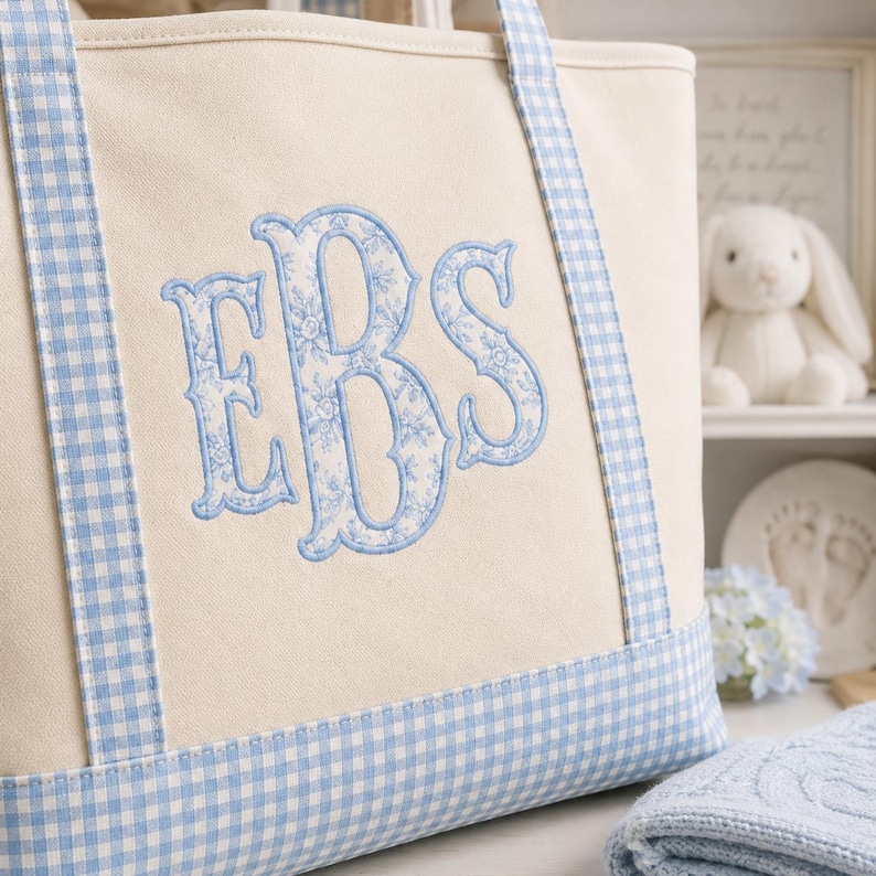 May include: A cream-coloured canvas tote bag featuring light blue gingham trim on the handles and base. The centre of the bag displays a large, embroidered monogram with the letters EBS in a light blue floral pattern with a solid blue border.