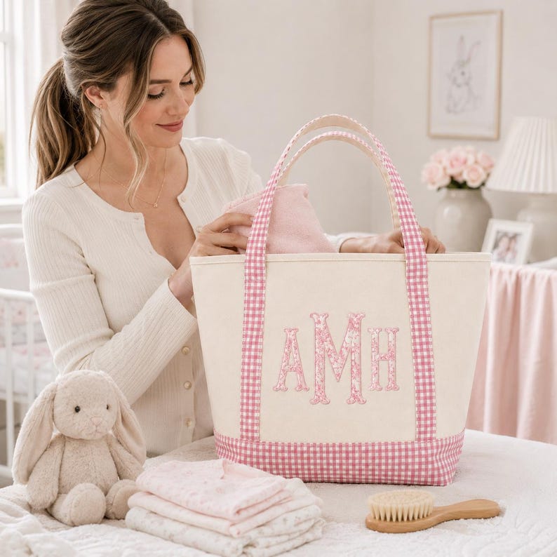 May include: A cream-coloured canvas tote bag featuring pink gingham trim on the handles and base. The front of the bag is embroidered with the pink floral monogram letters AMH. The bag is structured with two sturdy handles and a flat bottom base.