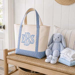 May include: A cream-colored canvas tote bag featuring blue and white gingham trim on the base and handles. The front of the bag displays a large, embroidered monogram with the letters EBS in a decorative blue font with a floral pattern.