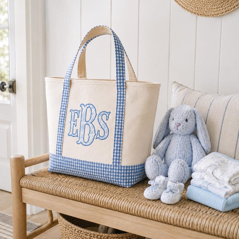 May include: A cream-colored canvas tote bag featuring blue and white gingham trim on the base and handles. The front of the bag displays a large, embroidered monogram with the letters EBS in a decorative blue font with a floral pattern.