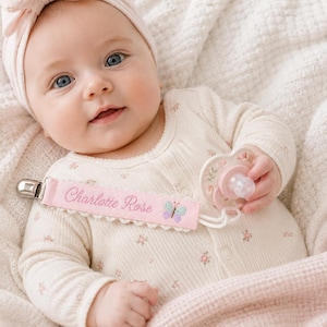 May include: A light pink pacifier clip featuring the embroidered name Charlotte Rose in cursive script and a small butterfly motif. The clip is attached to a pacifier with a floral pattern, worn by an infant in a cream-colored floral onesie.