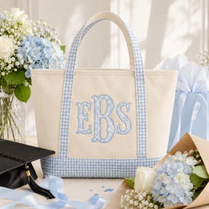 May include: A cream-coloured canvas tote bag featuring a light blue and white gingham pattern on the handles and base. The centre of the bag displays a large, embroidered monogram with the letters EBS in a light blue floral print fabric.