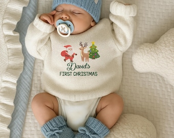 Op de afbeelding: Een baby met een lichtblauwe gebreide muts, bijpassende babyslofjes en een crèmekleurige trui met een kerstontwerp. De trui heeft de kerstman, een rendier en een kerstboom, met de tekst "David's FIRST CHRISTMAS". De baby draagt ook een fopspeen.