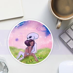 May include: Round sticker featuring an illustration of an alien embracing a person with dark skin, set against a dreamy, pastel-colored space scene. The sticker has the artist's signature, and the overall theme is one of love and connection.
