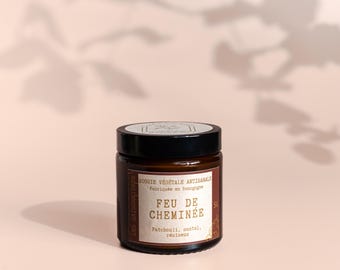 May include: A brown glass jar with a black lid, labelled "FEU DE CHEMINÉE", indicating a vegetable candle made in Burgundy, France. The label mentions patchouli, sandalwood, and resin. The jar is on a pink surface, with leaf shadows.