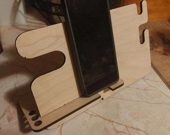 Op de afbeelding: Een houten telefoonstandaard met een zwarte telefoon in de houder. De standaard heeft een uitsparing voor een oplaadkabel en een klein gaatje voor een koptelefoonaansluiting.