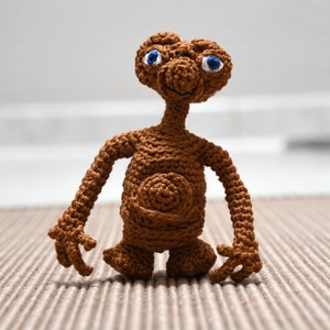 K&ouml;nnte beinhalten: Eine braune, geh&auml;kelte Figur der au&szlig;erirdischen Figur E.T. mit gro&szlig;en blauen Augen. Die handgefertigte Puppe hat einen langen Hals, Arme und Beine sowie eine detaillierte H&auml;kelstruktur. Die Figur steht auf einer neutralfarbenen Oberfl&auml;che.
