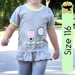 Puede incluir: Una camiseta gris con un diseño floral, con una flor rosa y un diseño circular blanco y negro. La camiseta tiene ribetes verdes en las mangas y el cuello, y un dobladillo con volantes. El texto "Size 116" está en una pancarta vertical verde.