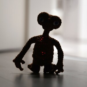 K&ouml;nnte beinhalten: Eine handgefertigte, geh&auml;kelte braune Alien-Figur mit gro&szlig;en Augen und einem langen Hals. Der Alien hat eine rot leuchtende Mitte und steht auf einer dunklen Oberfl&auml;che. Die Figur ist wahrscheinlich ein Spielzeug oder ein dekoratives Objekt.