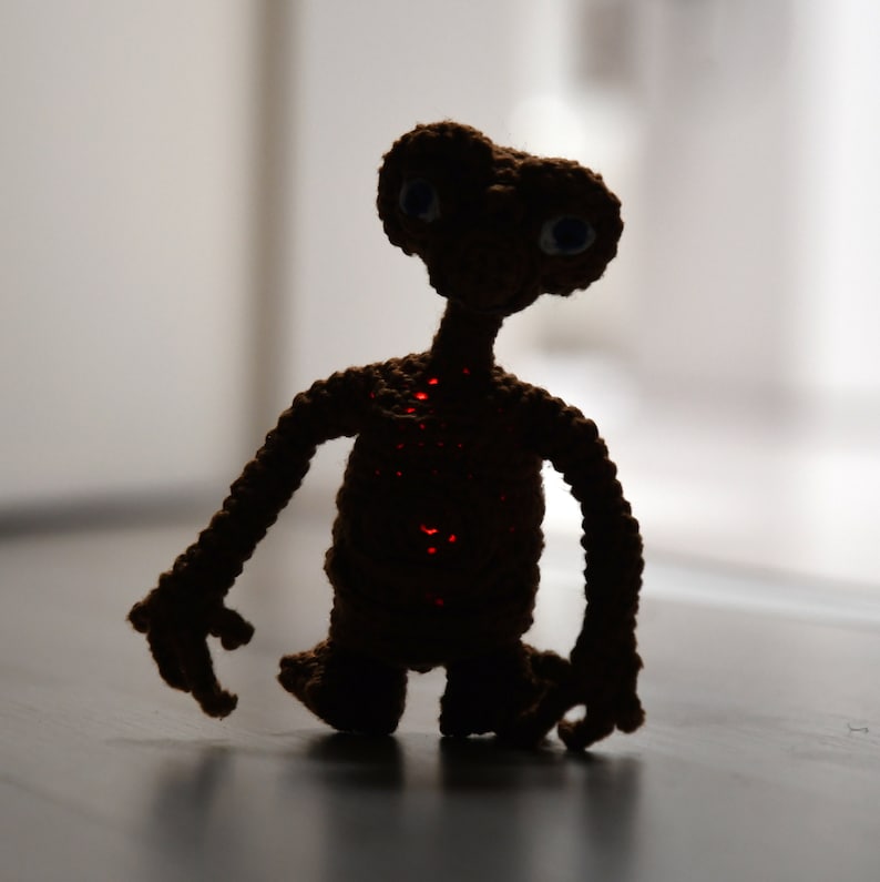 K&ouml;nnte beinhalten: Eine handgefertigte, geh&auml;kelte braune Alien-Figur mit gro&szlig;en Augen und einem langen Hals. Der Alien hat eine rot leuchtende Mitte und steht auf einer dunklen Oberfl&auml;che. Die Figur ist wahrscheinlich ein Spielzeug oder ein dekoratives Objekt.