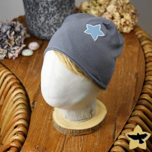 Può includere: Un berretto grigio con un'applicazione a stella azzurra. Il cappello è esposto su una testa di manichino sopra una fetta di legno. Lo sfondo include fiori secchi e un vaso decorativo.