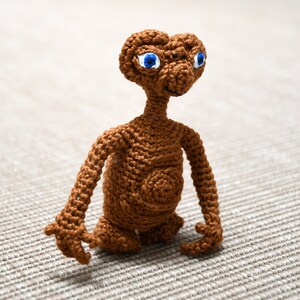 K&ouml;nnte beinhalten: Eine handgefertigte, braune H&auml;kelfigur des Au&szlig;erirdischen E.T. aus dem Film E.T. Der Au&szlig;erirdische. Die Figur hat gro&szlig;e blaue Augen, einen langen Hals und ausgestreckte Arme. Die H&auml;kelarbeit ist detailliert.
