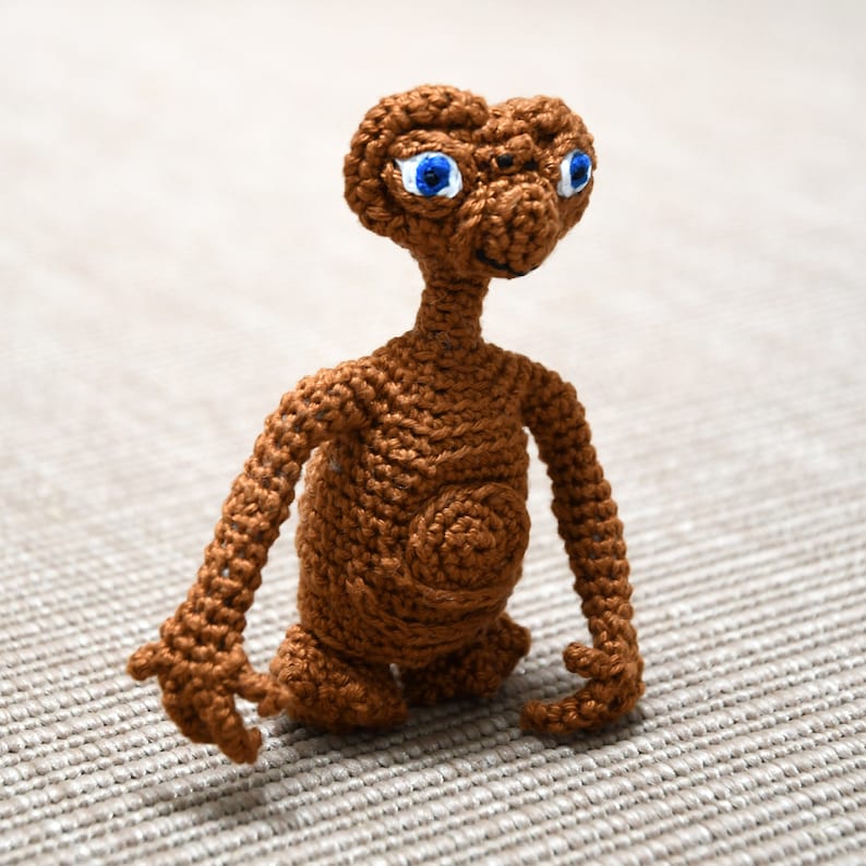 K&ouml;nnte beinhalten: Eine handgefertigte, braune H&auml;kelfigur des Au&szlig;erirdischen E.T. aus dem Film E.T. Der Au&szlig;erirdische. Die Figur hat gro&szlig;e blaue Augen, einen langen Hals und ausgestreckte Arme. Die H&auml;kelarbeit ist detailliert.