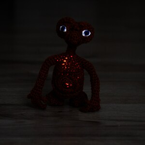 K&ouml;nnte beinhalten: Eine handgefertigte, geh&auml;kelte Figur von E.T. dem Au&szlig;erirdischen. Der r&ouml;tlich-braune Au&szlig;erirdische hat gro&szlig;e, blau&auml;ugige Augen und leuchtend orangefarbene Lichter auf seinem K&ouml;rper. Die Figur steht auf einer Holzoberfl&auml;che.