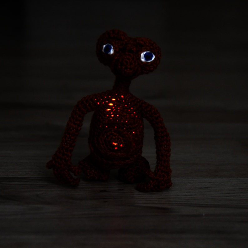 K&ouml;nnte beinhalten: Eine handgefertigte, geh&auml;kelte Figur von E.T. dem Au&szlig;erirdischen. Der r&ouml;tlich-braune Au&szlig;erirdische hat gro&szlig;e, blau&auml;ugige Augen und leuchtend orangefarbene Lichter auf seinem K&ouml;rper. Die Figur steht auf einer Holzoberfl&auml;che.
