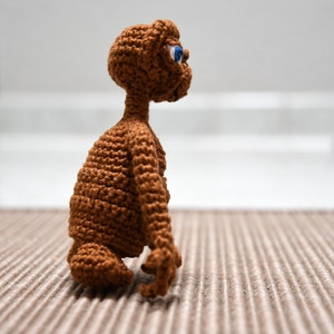 K&ouml;nnte beinhalten: Eine handgefertigte, braune E.T.-Figur aus H&auml;kelarbeit mit blauen Augen. Die Figur sitzt auf einer strukturierten Oberfl&auml;che und zeigt die detaillierte H&auml;kelarbeit. Arme und Kopf der Figur sind sichtbar und erfassen das Aussehen der ikonischen Figur.
