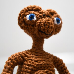 K&ouml;nnte beinhalten: Eine geh&auml;kelte braune Alien-Puppe mit gro&szlig;en blauen Augen und einem kleinen L&auml;cheln. Die handgefertigte Puppe weist detaillierte N&auml;hte und ein klassisches Charakterdesign auf, perfekt f&uuml;r Science-Fiction-Fans.